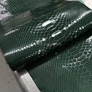 Puede incluir: Una lámina de cuero de piel de serpiente verde oscuro con un acabado brillante. El cuero está enrollado, revelando el patrón texturizado de las escamas. Las escamas son hexagonales y están dispuestas en un patrón repetitivo.
