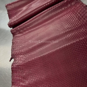 Può includere: Primo piano di pelle bordeaux con un motivo strutturato a scaglie. La pelle è piegata e sembra essere in fogli. Il materiale ha una leggera lucentezza, che suggerisce una finitura lucida. I bordi sono irregolari.