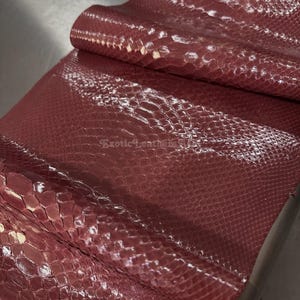Könnte beinhalten: Nahaufnahme einer Rolle exotischen Leders in Burgunderrot. Das Leder hat ein strukturiertes, schuppenartiges Muster und eine glänzende Oberfläche. Die Worte "Exotic Leather" sind in der unteren rechten Ecke zu sehen.