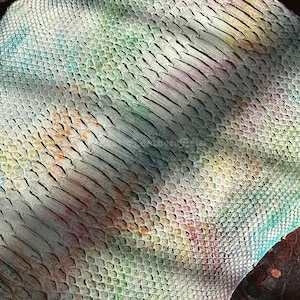 May include: Close-up of a piece of leather with a scale pattern. The leather is dyed with a watercolor effect, featuring pastel shades of pink, yellow, green, and blue. The scales have a textured appearance.