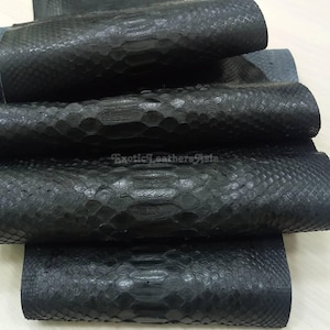 May include: Close-up of several rolls of black snakeskin leather. The leather has a textured, scaly pattern and a matte finish. The rolls are stacked on top of each other, showcasing the material's natural texture and color.
