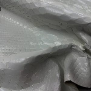 Könnte beinhalten: Nahaufnahme einer weißen Schlangenhaut. Das Leder hat ein strukturiertes, schuppenartiges Muster und ist leicht zerknittert, wobei Glanzlichter und Schatten Tiefe verleihen. Das Material wirkt weich und geschmeidig.