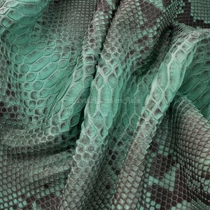 May include: Close-up of a folded piece of teal-colored snakeskin leather. The leather has a scale pattern with dark grey accents. The texture is visible, showing the natural details of the material. The overall appearance is luxurious and exotic.