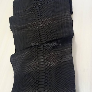 Puede incluir: Un trozo doblado de cuero de piel de serpiente negro. El cuero tiene un patrón texturizado y escamoso con un acabado oscuro y mate. El texto "Exotic Leathers Asia" es visible en la esquina inferior derecha.