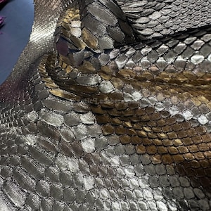 Puede incluir: Primer plano de cuero de serpiente plateado y dorado metálico. Las escamas tienen un patrón texturizado y superpuesto. El cuero está doblado, mostrando la parte inferior y resaltando las variaciones de color. El efecto general es lujoso y llamativo.