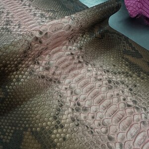 Può includere: Primo piano di un motivo a pelle di serpente. La pelle ha una miscela di toni grigi, marroni e rosa, con una consistenza squamosa. Le squame variano per dimensioni e forma, creando un design naturalistico. La pelle è piegata, mostrando il lato inferiore.