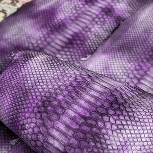 Può includere: Primo piano di diversi rotoli di pelle di serpente viola. La pelle ha un motivo a scaglie e un gradiente di tonalità viola, dal chiaro allo scuro. La trama è dettagliata e il materiale sembra di alta qualità.