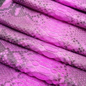 Può includere: Primo piano di pelle di serpente tinta impilata in tonalità rosa e grigio. Le squame sono visibili, creando una superficie strutturata. La pelle è piegata, mostrando il lato inferiore e i bordi del materiale.