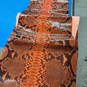 Puede incluir: Primer plano de un trozo de cuero de piel de serpiente naranja y negro. El cuero tiene una textura escamosa y un patrón natural. Los colores son vibrantes y el patrón es intrincado. La piel de serpiente se utiliza probablemente para accesorios de moda u otros artículos de cuero.