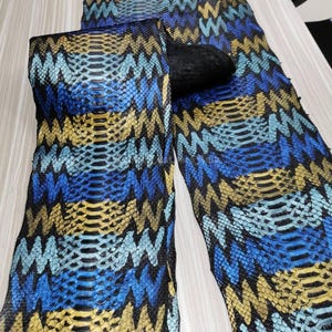 Può includere: Un tessuto a fantasia pelle di serpente nei toni del blu, giallo e nero. Il materiale è piegato, rivelando il disegno delle squame. Il motivo ha un design a zig-zag. Il tessuto sembra essere pelle o un materiale simile alla pelle.