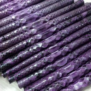May include: Close-up of several pieces of purple snakeskin leather. The leather has a textured, scaly pattern with dark grey and black accents. The pieces are arranged diagonally, showcasing the natural texture and color variations.