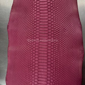 Puede incluir: Primer plano de un trozo de cuero de serpiente de color burdeos. El cuero tiene un patrón texturizado y escamoso con una columna vertebral más oscura en el centro. Las escamas son pequeñas y están muy juntas.