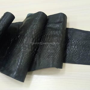 May include: Close-up of a rolled piece of black snakeskin leather. The leather has a textured, scaly pattern and is folded over itself. The text "Exotic Leathers Asia" is visible in the image.