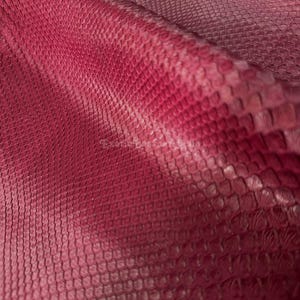 Puede incluir: Primer plano de una hoja de cuero de piel de serpiente roja y rosa. El cuero tiene un patrón texturizado en forma de escamas. El color pasa de un rojo intenso a un tono rosa más claro.