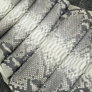 Può includere: Primo piano di diversi pezzi di pelle di serpente grigia e bianca. Le pelli sono piegate e stratificate, rivelando gli intricati motivi di squame e le trame. La tavolozza dei colori è principalmente neutra, con variazioni di grigio e bianco.