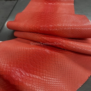 Può includere: Un foglio di pelle rosso vibrante piegato, con un motivo a squame di rettile. Il materiale ha una finitura lucida e viene probabilmente utilizzato per l'artigianato o la moda. L'immagine è ben illuminata, evidenziando la consistenza e il colore della pelle.