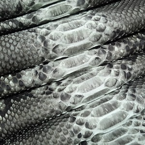 Può includere: Primo piano di pelli di cuoio impilate e strutturate in tonalità grigie e bianche. Le pelli presentano un motivo a scaglie, con un mix di toni scuri e chiari che creano un aspetto dettagliato e naturalistico.