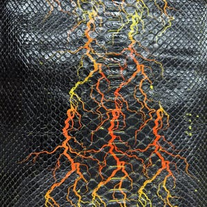 May include: Close-up of a textured black surface with a scale-like pattern. Bright orange and yellow lightning bolt-like designs are painted across the surface, creating a striking contrast. The design resembles reptile skin.
