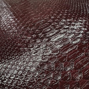 May include: A close-up shot of a sheet of burgundy-colored leather with a reptile scale pattern. The leather has a glossy finish, reflecting light across the surface. The scales are tightly packed, creating a textured appearance.