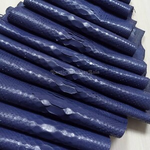 May include: A collection of rolled navy blue snakeskin leather pieces. The leather displays a textured, scale-like pattern and a shiny surface. The image includes the text "Exotic Leathers Asia".