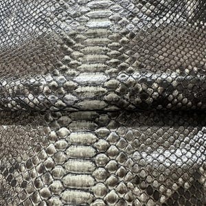 Può includere: Primo piano di un motivo a pelle di serpente. Le squame sono un mix di grigio, argento e nero, creando una superficie strutturata. Il motivo è simmetrico, con un design centrale a forma di spina dorsale e squame a forma di diamante.