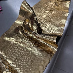May include: Close-up of a sheet of gold-colored snakeskin leather. The leather has a textured, scale-like pattern and is folded over itself. The metallic sheen reflects light, highlighting the intricate details of the material.