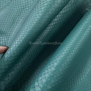 Può includere: Primo piano di pelle di serpente color verde acqua. La pelle ha un motivo squamoso strutturato ed è piegata, rivelando il lato inferiore. Il testo "Exotic Leathers Asia" è visibile nell'angolo in basso a destra.