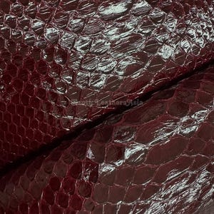 Può includere: Primo piano di un foglio di pelle bordeaux con finitura lucida e strutturata. La pelle presenta un motivo a scaglie, creando un aspetto di pelle di rettile. La superficie riflette la luce, evidenziando la consistenza e le variazioni di colore.