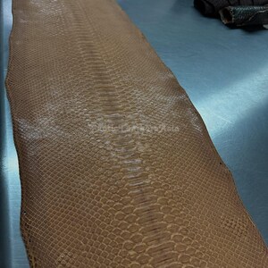 Puede incluir: Una gran pieza de cuero de piel de serpiente marrón con un patrón de escamas. El cuero es de color marrón claro y parece flexible. La piel de serpiente está sobre una superficie metálica.