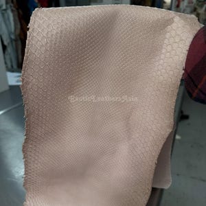 Puede incluir: Un trozo de cuero de piel de serpiente de color beige claro con un patrón de escamas. El cuero se sostiene, mostrando su textura y color. La imagen está tomada en un taller.