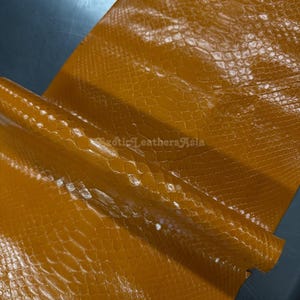 May include: A close-up shot of a sheet of tan-coloured exotic leather with a snakeskin pattern. The leather has a glossy finish and is folded over. The text "Exotic Leathers Asia" is visible.