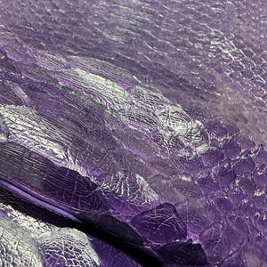 May include: Close-up of a textured, purple material with a scale-like pattern. The surface has a glossy finish, with highlights and shadows creating depth. The material appears to be leather or a similar fabric.