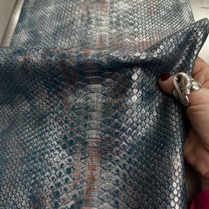 Puede incluir: Primer plano de un trozo de cuero sintético con un patrón de piel de serpiente. El material tiene una combinación de colores verde azulado y plateado con acentos marrones. Las escamas son detalladas y texturizadas, creando una apariencia realista. El material está doblado, mostrando su flexibilidad.