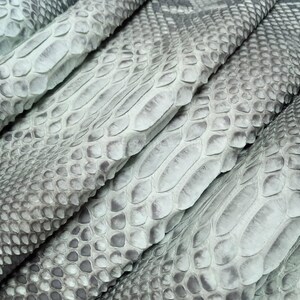 以下が含まれることがあります： 薄い灰色のヘビ革のシートのクローズアップ。 うろこが見え、質感のあるうろこ模様です。 革は折りたたまれ、重ねられており、奥行きと質感が生まれています。 全体的な色合いは落ち着いており、グレーと白のトーンです。