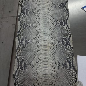 Può includere: Una pelle di serpente con un motivo di squame grigie, nere e bianche. La pelle è rettangolare e sembra appena conciata, con una consistenza morbida. Le squame sono dettagliate e creano un design naturalistico.