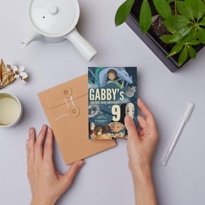K&ouml;nnte beinhalten: Eine Person h&auml;lt eine farbenfrohe Einladungskarte mit dem Text "Gabby's Spirited Away Adventure 9". Die Karte zeigt Illustrationen aus dem Animationsfilm. Ein brauner Umschlag, eine wei&szlig;e Teekanne und eine Tasse Tee sind ebenfalls zu sehen.