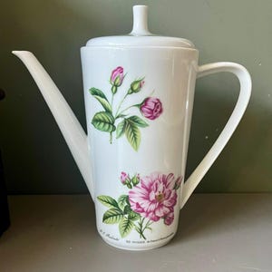 Peut inclure: Une cafetière en porcelaine blanche avec un corps cylindrique, un couvercle à bouton, une anse incurvée et un long bec. La surface présente des illustrations botaniques de roses roses et de feuilles vertes, avec le texte P.J. Redouté XII ROSIER de France.