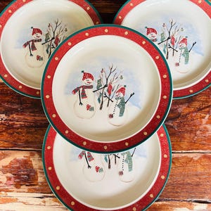 Puede incluir: Cuatro platos redondos de cerámica con una escena invernal festiva que muestra dos muñecos de nieve con bufandas y gorros rojos y verdes, junto a un árbol desnudo decorado con calcetines. Los platos tienen un borde rojo con puntos dorados.