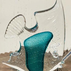 Op de afbeelding: Een heldere glazen vissculptuur met turquoise accenten. De sculptuur heeft een getextureerd turquoise gedeelte en is gevuld met kleine belletjes. De vis heeft een vin aan de bovenkant en een staartvin. De sculptuur staat op een bruin, getextureerd oppervlak.
