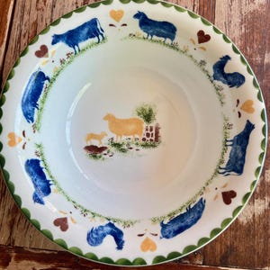 Può includere: Ciotola in ceramica bianca con bordo smerlato verde. La ciotola presenta mucche blu, pecore e un gallo, insieme ad accenti a cuore gialli e marroni. Una scena pastorale con una pecora e un agnello è al centro.
