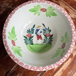 Op de afbeelding: Een ronde keramische kom met een witte basis en een handgeschilderde centrale illustratie van een persoon die doedelzak speelt. De binnenzijde is versierd met groene bladmotieven, rode bloemen en een herhalend rood schulprandpatroon.