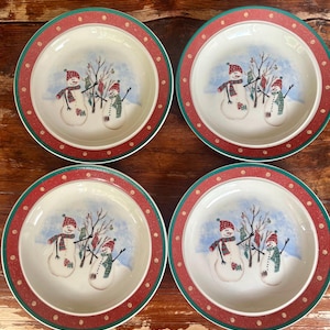 Puede incluir: Un juego de cuatro platos redondos de cerámica con una ilustración central de dos muñecos de nieve junto a un árbol desnudo. Los platos tienen un borde ancho rojo decorado con pequeños puntos blancos y un borde exterior verde.