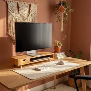 May include: A modern home office setup featuring a wooden desk with a monitor, keyboard, and mouse. A wooden monitor stand with a drawer sits on the desk. A macrame wall hanging and potted plants add decorative elements.