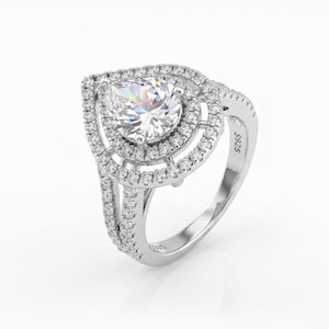 May include: A silver ring with a large, pear-shaped, clear gemstone at the center, surrounded by two halos of smaller, clear stones. The band is also adorned with small, clear stones. The ring is set against a white background.