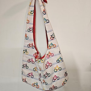 May include: White shoulder bag with a red interior and strap. The bag is patterned with colorful bicycles and blue horizontal stripes. The bag is hanging against a white wall.