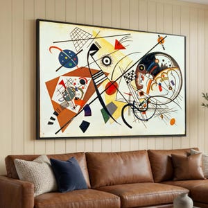 Puede incluir: Impresión de arte abstracto en un marco negro. La obra presenta formas geométricas, líneas y círculos en una variedad de colores, incluyendo rojo, amarillo, azul y negro, sobre un fondo blanco. La pintura se exhibe sobre un sofá de cuero marrón.