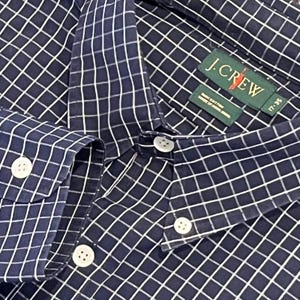 May include: A navy blue long-sleeve button-down shirt featuring a white grid pattern. The shirt has a pointed collar, white buttons, and a green J.Crew brand label on the inside neck area with the text 17-35 and 100% cotton visible.