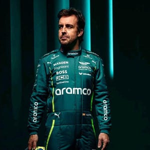 May include: A teal racing suit with multiple sponsor logos, including "aramco," "MAADEN," and "BOSS." The suit has yellow accents and the name "ALONSO" is visible. The person is looking off to the side against a dark green background.