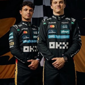 May include: Two people in black racing suits, featuring sponsor logos such as "Android" and "Dell Technologies." The suits have white and yellow detailing. A checkered flag is visible in the background.