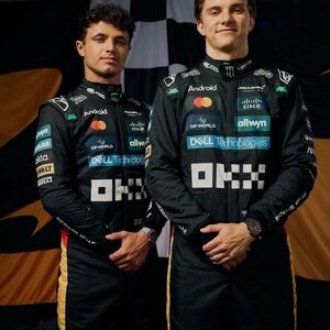 May include: Two people in black racing suits with sponsor logos, including "Android," "Dell Technologies," and "Cisco." The suits have white and yellow accents. They are standing in front of a checkered flag background.